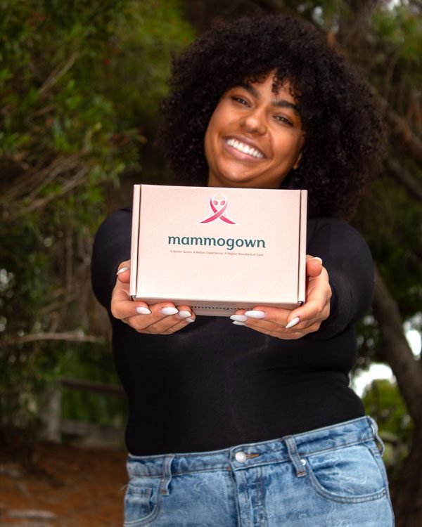 Woman outdoors holding MammoGown gift box toward camera.